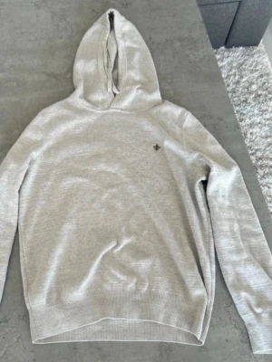 Ljusgrå hoodie med broderad detalj morris  - Ljusgrå hoodie med huva och långärmad design. Materialet är stickat och mjukt, perfekt för en avslappnad look. På bröstet finns en liten broderad fleur-de-lis i mörk färg som ger en stilren touch. Ribbstickade muddar vid ärmslut och nederkant.