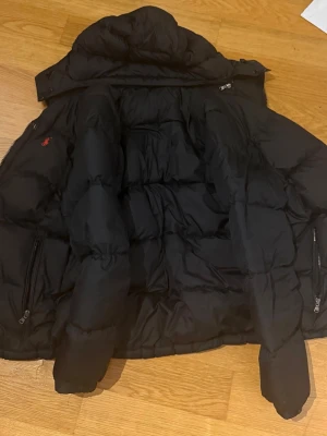 Svart pufferjacka från Polo Ralph Lauren - Svart pufferjacka från Polo Ralph Lauren med långärmad design och quiltad struktur. Jackan har dragkedjor på sidorna och en liten röd logotyp på bröstet. Perfekt för kalla dagar och har en hög krage för extra värme. Med avtagbar luva 