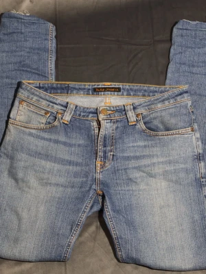 Nudie jeans  - Dessa Nudie Jeans är tillverkade av 100 % ekologisk bomull, med en klassisk ljusblå tvätt med naturliga morrhår och märkets signaturorangea sömmar på bakfickorna. Den smala passformen ger en tidlös look, perfekt för vardagsbruk.  ⭐️ Skick: 7/10 (Mindre slitage i linje med regelbunden användning, lätt fransning vid fållen) 🎲 Storlek: W 29 L 32 🎨 Färg: Ljusblå 📦 Snabb och noggrann leverans.