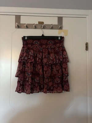 Mönstrad volangkjol från Lindex - Säljer en snygg kort volangkjol från Lindex i storlek 158/164 (men passar mig som vanligtvis har S/XS). Kjolen är vinröd med fint mönster i blått, vitt och orange. Den har flera lager volanger och resår i midjan för en bekväm passform. Jättefin och passar perfekt till sommaren!