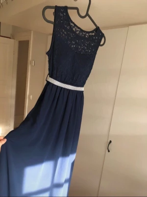 Marinblå långklänning med spets och glitter  - Elegant marinblå långklänning med spets upptill och ett glittrigt bälte i midjan samt knyte på ryggen. Klänningen är i ett lätt och bekvämt material med en tunn underkjol så att klänningen är inte genomskinlig, perfekt för speciella tillfällen. 