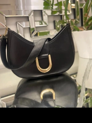 Svart axelväska med gulddetaljer - Stilren svart handväska i läderimitation med bred axelrem och snyggt guldfärgat U-spänne framtill. Väskan har en halvmåneform och detaljer i metall, perfekt för att lyfta din outfit.