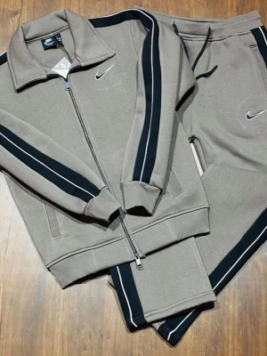 Grå mjukisbyxor från Nike med svarta ränder - Snygga grå mjukisbyxor från Nike med svarta och vita ränder längs sidorna. Byxorna har dragsko i midjan och är tillverkade i mjukt material. Perfekta för en avslappnad stil och matchar med tillhörande ziptröja.