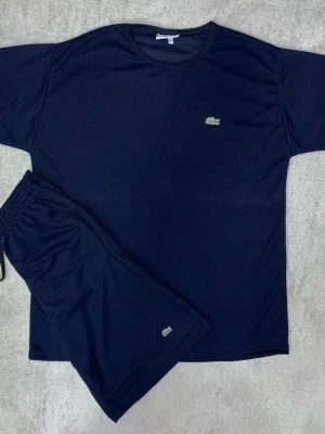 Mörkblå Lacoste set med t-shirt och shorts - Snyggt mörkblått set från Lacoste med en enkel t-shirt och matchande shorts. Båda plaggen har den klassiska Lacoste-loggan broderad framtill. Materialet känns mjukt och bekvämt, perfekt för chill dagar. Tidlös design med rund hals och elastisk midja på shortsen.