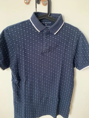 Mörkblå prickig pikétröja Primark XS - Snygg mörkblå pikétröja från Primark i storlek XS. Tröjan har vita små prickar över hela, klassisk krage med vit kant och tre knappar framtill. Perfekt för en stilren och avslappnad look.