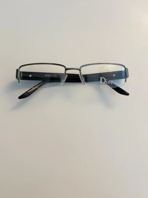 Svarta glasögonbågar från Dior - Stilrena glasögonbågar från Dior i svart och silver med rektangulär form. Bågarna har diskret Dior-logga på sidan och näskuddar för extra komfort. Perfekt accessoar för dig som vill ha en klassisk och lyxig look.