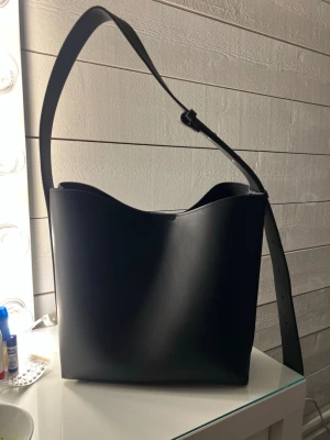 Svart axelväska i läderlook - Stilren svart axelväska med justerbart axelband och minimalistisk design. Väskan har en rymlig insida med innerficka som stängs med dragkedja och magnetlås upptill. Perfekt för dig som gillar enkel och modern stil.