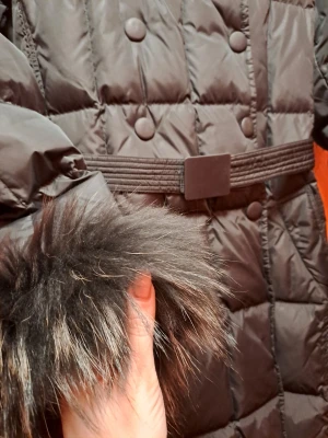 Svart lång dunjacka med äkta päls - Snygg svart lång dunjacka med markerad midja och bälte. Jackan har stora knappar framtill och är dekorerad med äkta päls vid krage och ärmslut. Perfekt för kalla vinterdagar och ger en trendig look.