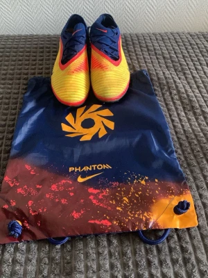 Nike Phantom fotbollsskor gul/röd/blå - Säljer ett par Nike Phantom Haaland personal edition. Skorna är endast använda ett fåtal gånger och har fortfarande en skinande Nike logga. Säljer de pga de vart för stora för mig. Påsen följer med och är i helt nyskick. Skorna har en ag platta för konstgräs spel.