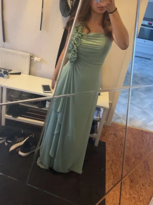 Ljusgrön långklänning med blommor - Elegant ljusgrön långklänning med ett axelband och vackra blommiga detaljer på axeln. Klänningen har draperad överdel och faller mjukt i flera lager. Perfekt för speciella tillfällen där du vill sticka ut.