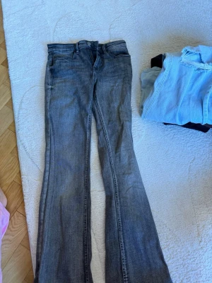 Grå bootcut jeans från Vero Moda - Säljer ett par grå bootcut jeans från Vero Moda. Jeansen har klassisk femficksdesign, knappstängning och är tillverkade i denim. Snyggt slitna detaljer och en cool vintage-look. Perfekta för dig som gillar en avslappnad och trendig stil.