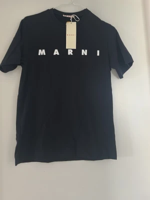 Svart Marni t-shirt med logga - Snygg svart t-shirt från Marni med vit logga tryckt över bröstet. Klassisk rund halsringning och korta ärmar. Tillverkad i mjuk bomull för en skön känsla och enkel stil. Perfekt för dig som gillar minimalistisk design och streetwear-vibe. den är aldrig använd hör av dig vid frågor Pris går att diskutera.