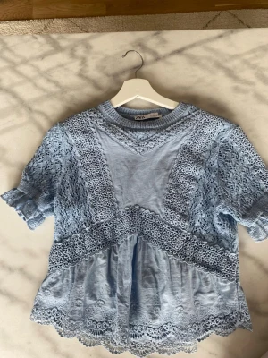 Ljusblå spetsblus från Zara - Supersöt ljusblå blus från Zara med detaljerad spets och broderade kanter. Blusen har korta ärmar, rund halsringning och volang längst ner. Perfekt för dig som gillar romantiska och feminina plagg.