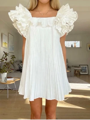 Vit plisserad klänning från Zara - Superfin vit kortklänning från Zara med plisserat tyg och stora volangärmar. Klänningen har en rak passform och fyrkantig ringning, vilket ger en trendig och luftig look. Perfekt för dig som gillar romantiska detaljer och en stilren vibe.