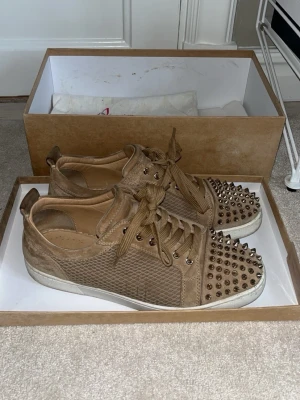 Beige Christian Louboutin sneakers med nitar - Snygga beige sneakers från Christian Louboutin med textur i mocka och läder. Skorna har coola metalnitar på tån och matchande beige snörning. Vit sula och exklusiv design som sticker ut. Perfekta för dig som vill ha något unikt och trendigt.