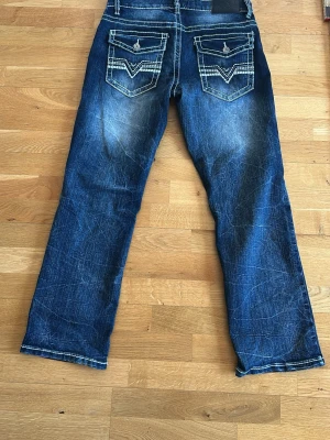 Blå true luck jeans med kontrastsömmar - Snygga blå jeans med tydliga vita kontrastsömmar och slitningar. Jeansen har raka ben och två bakfickor med lock och dekorativa sömmar. Perfekta för en avslappnad och trendig look.