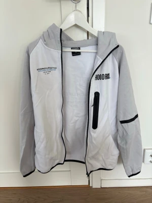 Hoodrich vit och grå hoodie med dragkedja - Vit och ljusgrå hoodie från Hoodrich med svart tryck på bröstet, ryggen och huvan. Dragkedja framtill, svart bröstficka med zip och svarta detaljer på ärmarna. Mjukt material och avslappnad passform för en modern streetstyle. Nypris ca 1000kr. Pris kan diskuteras vid snabb affär.