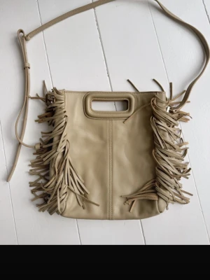 Maje väska helt oanvänd  - Cool beige maje axelväska i mjukt material med långa fransar på sidorna och ett praktiskt handtag upptill. Väskan har justerbar axelrem och en stilren, fyrkantig form som ger en trendig boho-vibe.