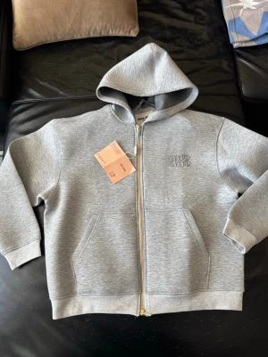 Grå hoodie med dragkedja från MiuMiu - Snygg grå hoodie från MiuMiu med dragkedja framtill och broderad logga på bröstet. Tröjan har huva, två fickor och ribbade muddar vid ärmslut och nederkant. Perfekt för en avslappnad och trendig look.