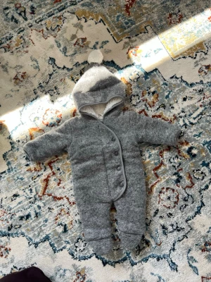 Grå jumpsuit med huva och knappar - Mysig grå jumpsuit för baby med huva och söt tofs upptill. Utsidan är i mjukt ull-liknande material och insidan är fodrad med vit fluffig fleece. Stängs med snedställda knappar framtill. Perfekt för kyliga dagar.
