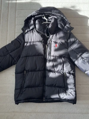 Svart pufferjacka från Polo Ralph Lauren - Snygg svart pufferjacka från Polo Ralph Lauren med röd logga på bröstet. Jackan har huva, två dragkedjeförsedda fickor och quiltad design. Perfekt för kalla dagar och har en sportig vibe. Materialet är glansigt och känns tåligt.