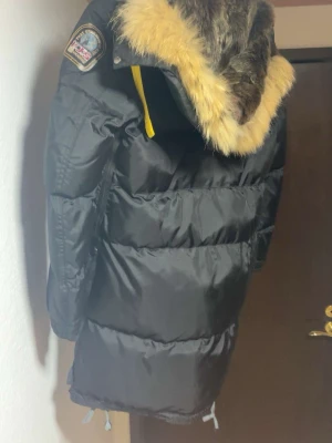 Svart dunjacka Parajumpers med päls - Säljer en svart dunjacka från Parajumpers med stor huva och fluffig pälsdetalj. Jackan har patch på ärmen, gula detaljer och är lång i modellen. Perfekt för kalla vinterdagar när du vill hålla dig varm och snygg.