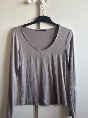 Beige långärmad topp från Vila - En enkel och stilren beige långärmad topp från Vila med rundad halsringning. Tillverkad i mjukt material som känns skönt mot huden. Perfekt att matcha med jeans eller kjol för en avslappnad look.