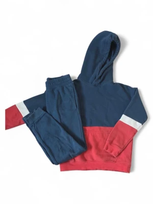 Blå och röd hoodie med vit detalj 146 - Snygg hoodie i färgblockad design med mörkblå överdel, röd nederdel och vita ränder på ärmarna. Stor känguruficka framtill och klassisk huva. Perfekt för en chill och avslappnad stil. Mjuk bomullsblandning som känns skön mot huden. Matchande mjukisbyxor. 