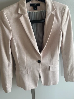 Rosa kavaj från H&M - Stilren beige kavaj från H&M med klassisk krage och en knapp framtill. Kavajen har två fickor med lock och är helfodrad med randigt innerfoder. Figurnära passform och långa ärmar, perfekt för en snygg och clean look.