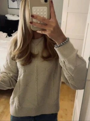 Beige stickad tröja med mönster - Mysig beige stickad tröja med diskret mönster och rund hals. Tröjan har långa ärmar och ribbade muddar vid ärmslut och nederkant. Perfekt för lager-på-lager och passar till jeans eller kjol.
