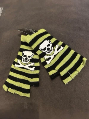 Randiga fingerlösa vantar med dödskalle - Fingerlösa vantar med svart och limegrön randigt mönster och en vit dödskalle med korslagda ben på ovansidan. Perfekta för dig som vill ha en alternativ stil och ändå hålla händerna varma.