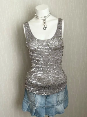 Glittrig silverfärgad paljettopp - Snygg ärmlös topp i silver med massor av paljetter som ger ett riktigt glittrigt intryck. Modellen är figurnära med breda axelband och rundad halsringning. Perfekt för dig som vill sticka ut och glänsa.