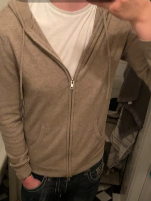 Beige cashmere hoodie från John Henric - Mjuk och lyxig beige hoodie i cashmere från John Henric. Tröjan har dragkedja framtill, två fickor och huva. Perfekt för en chill och stilren look. Nypris 2999kr