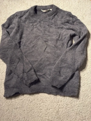 Mjuk grå fluffig stickad tröja - Supermjuk och fluffig stickad tröja i grått med rund hals och långa ärmar. Endast använd 1 gång! Perfekt för dig som gillar en cozy vibe och vill ha något riktigt gosigt att dra på sig. Tröjan har en enkel design utan synliga detaljer eller mönster.