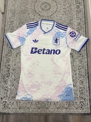 Aston Villa x Adidas retro matchtröja - Säljer en limited edition Aston Villa matchtröja från Adidas med retrodesign. Tröjan är vit med ljusrosa och ljusblå mönster, blå detaljer och klubbmärke på bröstet. Klassiska Adidas-ränder på axlarna och Betano-logga framtill. Perfekt för dig som älskar fotboll och unik stil.