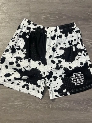 Svarta och vita meshshorts med tryck - Säljer ett par svarta och vita meshshorts från Eric Emanuel med coolt koskinnsmönster och logga på vänster ben. Shortsen har elastisk midja och är tillverkade i ett ventilerande meshmaterial som passar perfekt till sommaren.