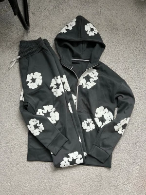 mörk grå hoodie och byxor från denim tears - Cool grå hoodie med dragkedja och stora vita blommor som mönster. Hoodien har huva och långa ärmar, perfekt för en avslappnad och trendig look. Materialet känns mjukt och skönt. Snygg detalj med hel dragkedja framtill.