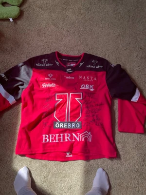 Örebro Hockey matchtröja signerad - Röd och svart matchtröja från Örebro Hockey med tryckta sponsorer och klubbens logga på bröstet. Tröjan är signerad av flera spelare och har långa ärmar. Perfekt för hockeyfans som vill sticka ut på läktaren.