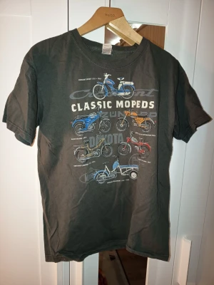 T-shirt med klassiska mopeder - Cool grå t-shirt med tryck av klassiska mopeder i olika färger och modeller. T-shirten har rund hals och korta ärmar. Perfekt för dig som gillar retro och mopeder.