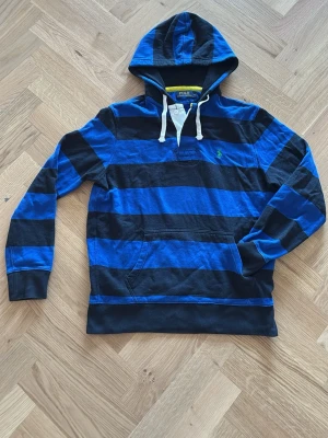 Randig hoodie från Polo Ralph Lauren - Snygg hoodie från Polo Ralph Lauren i blått och svart med breda ränder. Klassisk huva med vita snören, känguruficka och broderad logga på bröstet. Perfekt för en avslappnad och sportig stil.