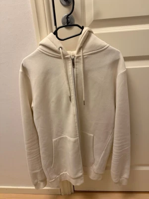 Vit zip-hoodie med fickor - Snygg vit hoodie med dragkedja framtill och två stora fickor. Klassisk modell med huva och dragsko. Perfekt för en avslappnad och stilren look. Mjuk och skön bomullskänsla.