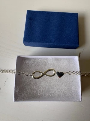 Silverfärgat armband med evighetstecken - Stilrent armband i silverfärg med dubbla kedjor, dekorerat med ett evighetstecken och ett litet hjärta. Perfekt accessoar för dig som gillar minimalistisk design och symbolik. Levereras i en blå ask. Stämpel 925 på armbandet (se bilden) 