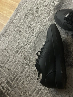 Svarta Nike Air Force 1 sneakers - Klassiska helsvarta Nike Air Force 1 sneakers i läder med låg profil och ikonisk swoosh på sidan. Skorna har en chunky sula och svarta skosnören, perfekt för en clean och stilren look.