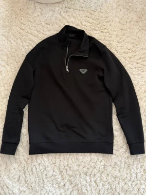 Svart prada half-zip hoodie  - Svart Prada hoodie med halv dragkedja och hög krage. På bröstet sitter en liten trekantig logga. Hoodien har långa ärmar och ribbade muddar. Perfekt för en clean och stilren look.