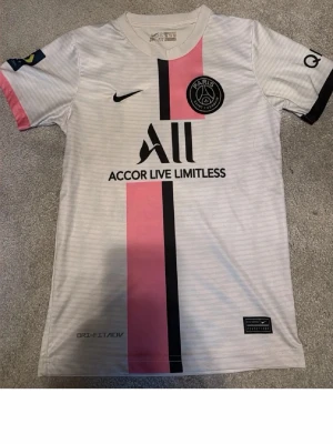 PSG Messi matchtröja vit/rosa - Säljer en PSG matchtröja från Nike med Messi och nummer 30 på ryggen. Tröjan är vit med tunna ränder, har en rosa och svart rand framtill samt PSG-logga på bröstet. Tillverkad i lätt funktionsmaterial, perfekt för fotboll eller träning.