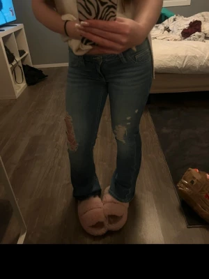 Blå slitna bootcut jeans - Säljer ett par blå bootcut jeans med slitningar på låren och knäna. Jeansen har klassisk femficksdesign och låg midja. Perfekta för en avslappnad och trendig look.