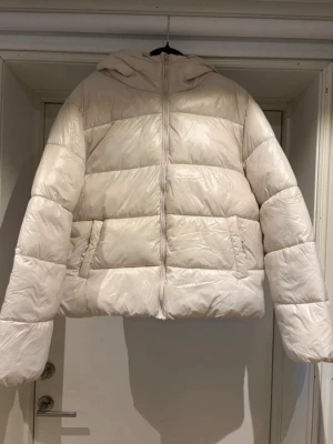 Vit pufferjacka med huva - Säljer en stilren vit pufferjacka med huva och dragkedja framtill. Jackan har breda, vadderade paneler och två fickor på sidorna. Perfekt för kalla vinterdagar och ger en riktigt cozy vibe. Materialet är glansigt och jackan har en boxig passform.