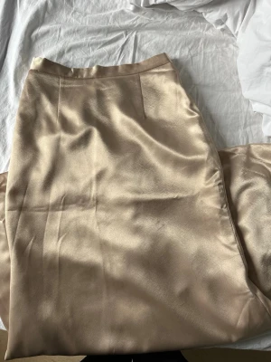Champagnefärgad satinkjol - Snygg långkjol i champagnefärgad satin med slät finish och enkel design. Kjolen har en rak passform.  Materialet ger ett lyxigt intryck. Dold dragkedja där bak. Högmidjad. Använts endast en gång. Finns tillhörnde korsett till kontakta för paketpris