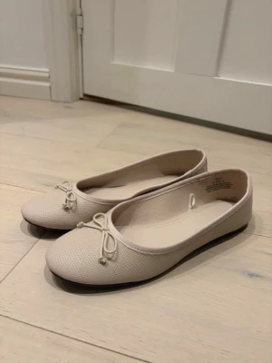 Beige ballerinaskor med rosett - Snygga beige ballerinaskor med rund tå och dekorativ rosett framtill. Skorna har en diskret textur i materialet och en tunn, platt sula. Perfekta för en stilren och enkel look.