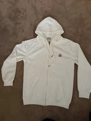 Vid Moncler Hoodie (storlek S) - Snygg vit hoodie från Moncler med dragkedja framtill och klassisk huva med dragsko. Tröjan har Moncler-logga på bröstet och praktiska fickor framtill. Perfekt för en clean och stilren look. Tillverkad i mjukt material för extra komfort. Säljs på grund av jag beställde fel storlek.
